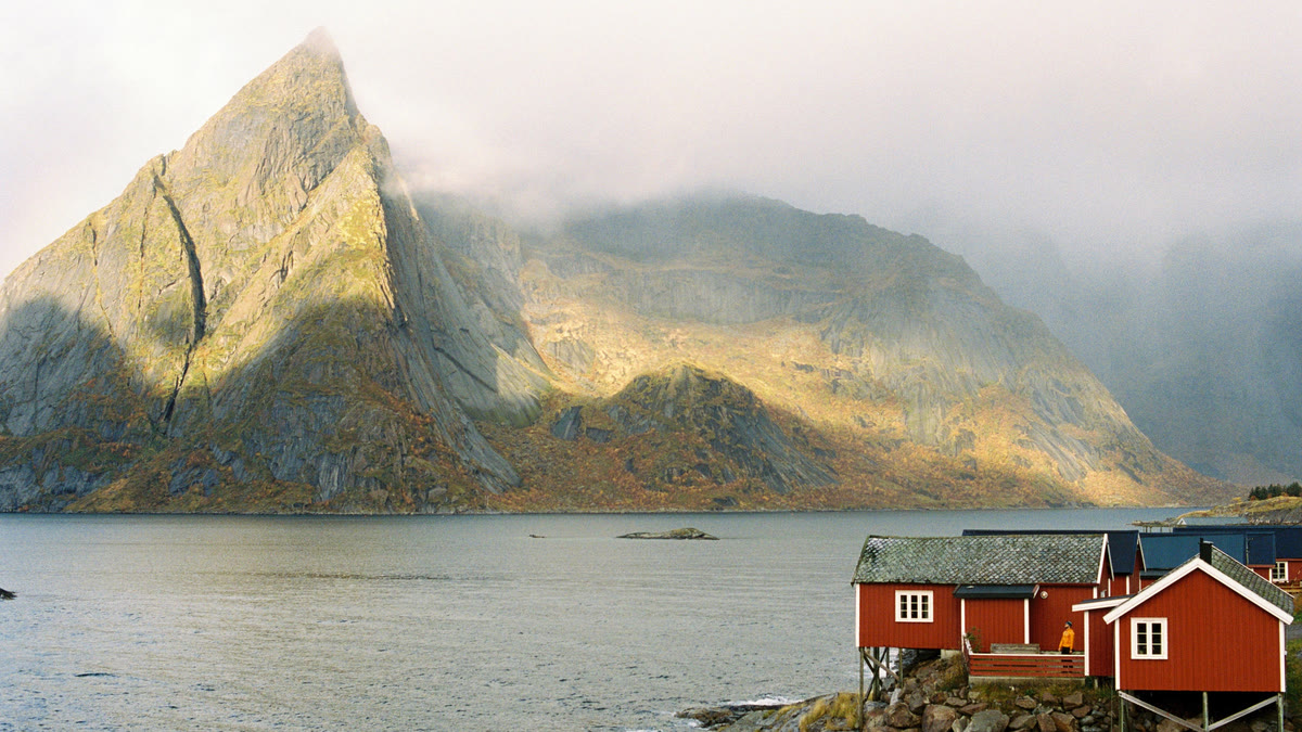 Red cabins on the shore beneath a sunlit, jagged peak
